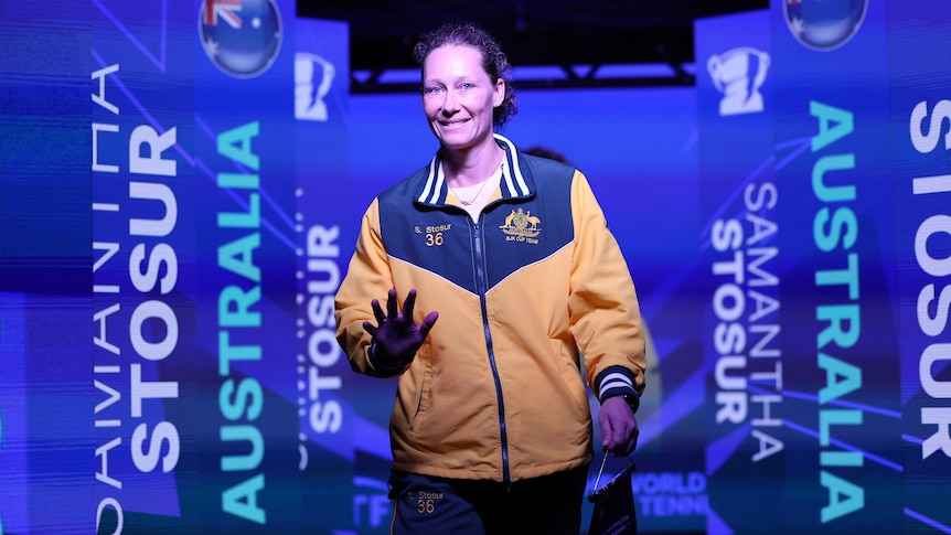 Samantha Stosur waves as she walks towards the camera while wearing green and gold Australia warm-up gear.
