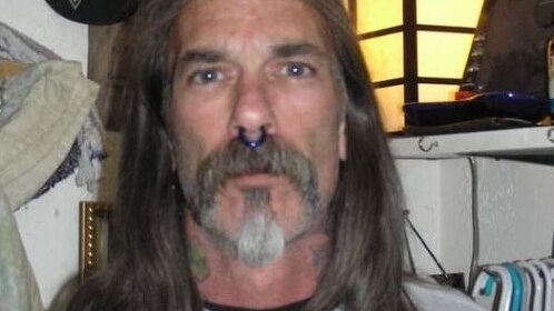 A man with a septum piercing, neck tattoos and long hair looks at the camera.