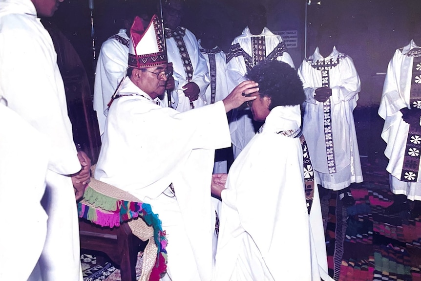 Reverend Sereima Lomaloma kneels in front of a seated bishop, who is marking her forehead with a cross.