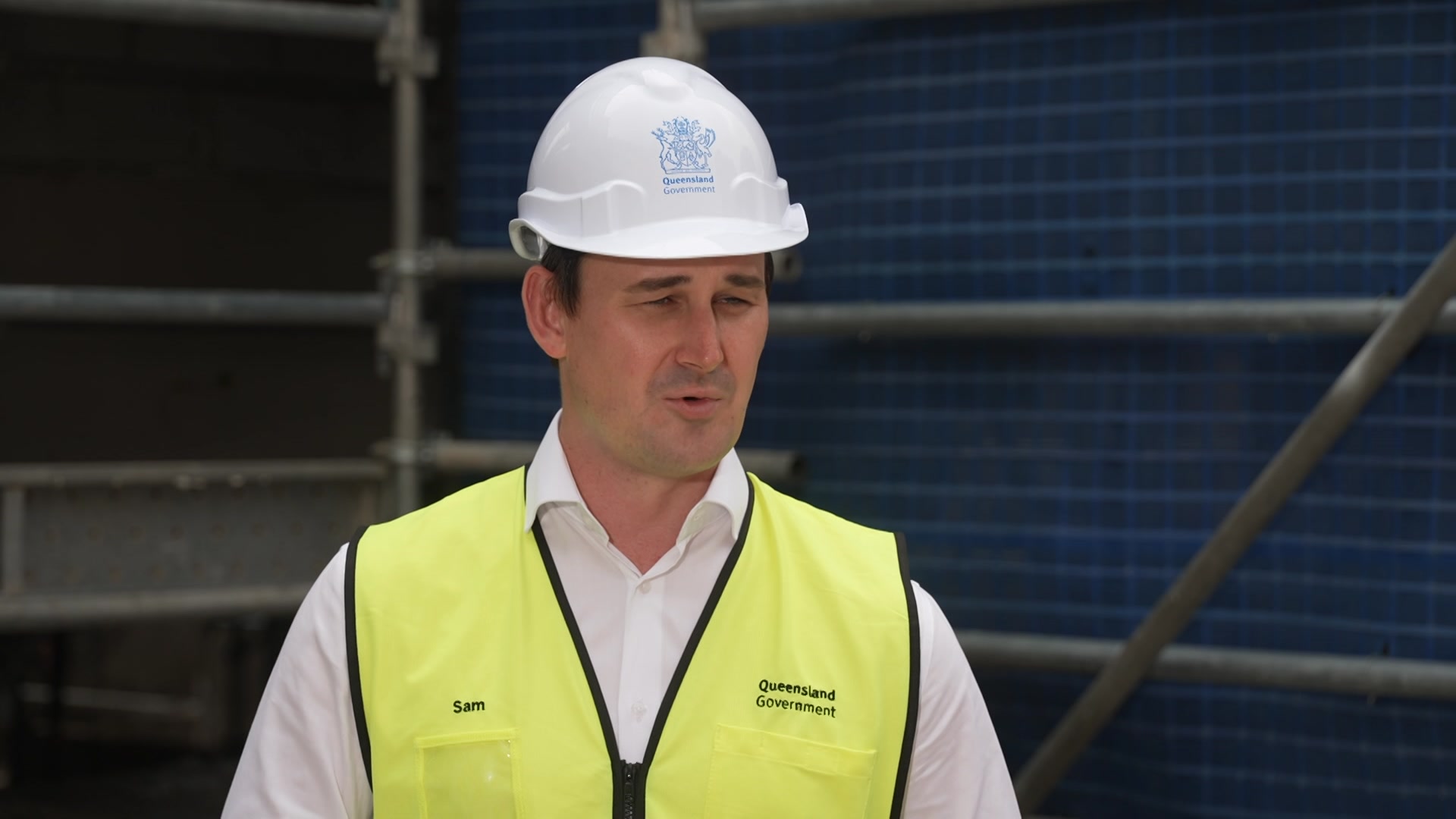 A man stands on a construction site wearing a hard hat and fluro yellow vest over a white shirt.