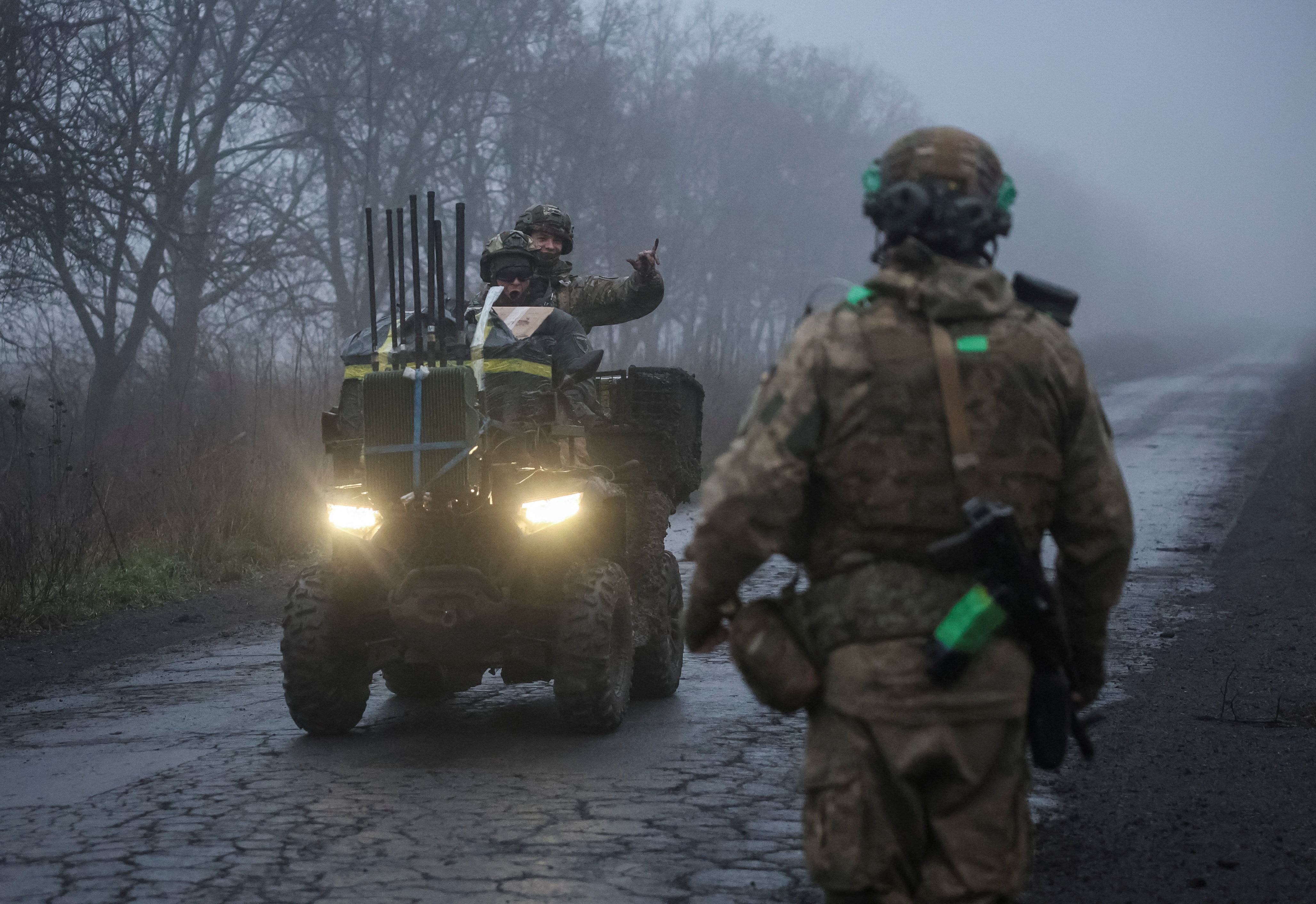 Two men in Ukrainian military gear riding on a buggy past another soldier on a misty road