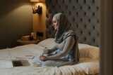 A woman wearing a hijab sitting on a bed with a laptop and holding a mug.