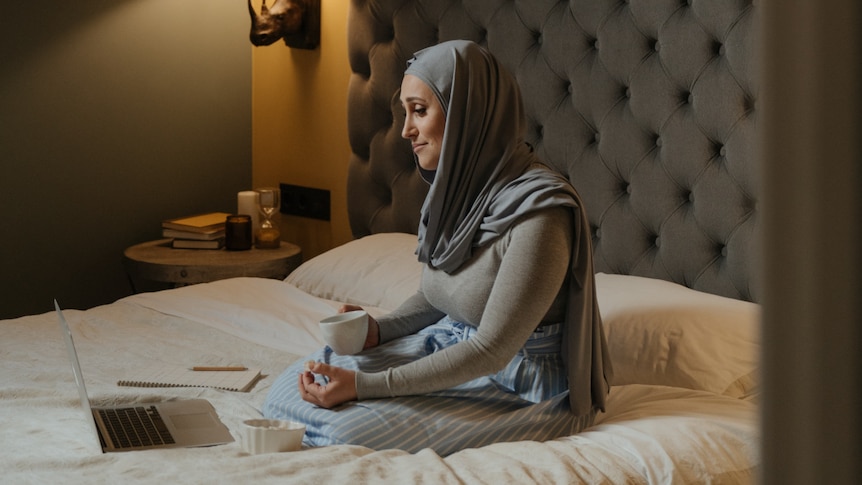 A woman wearing a hijab sitting on a bed with a laptop and holding a mug.