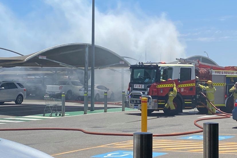 A fire engine, with fire fighters wearing yellow suits is in front of a large, black smoke cloud in a car park.