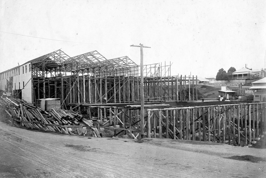 A black and white photo of a half-built timber shed on the side of a hill.