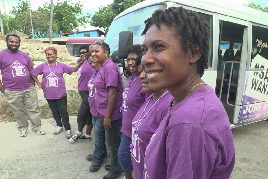 Seven Papua New Guineans stand in a semi-circle smiling in front of a bus.