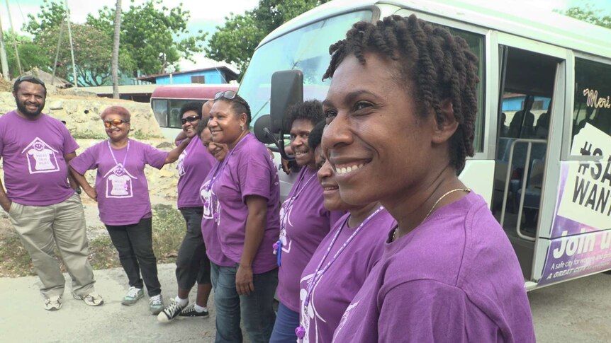 Seven Papua New Guineans stand in a semi-circle smiling in front of a bus.