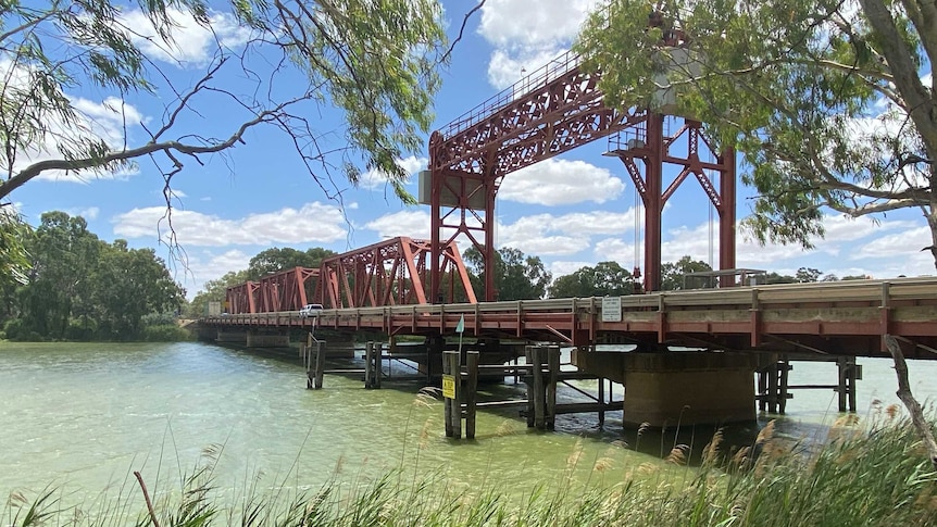 A large, red bridge spans a river while trees and reeds appear in the foreground.