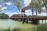 A large, red bridge spans a river while trees and reeds appear in the foreground.