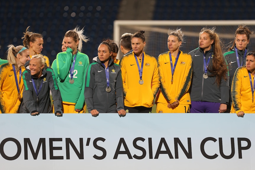 A group of Australia's Matildas players stand behind a sign for the Women's Asian Cup wearing their runners-up medals.