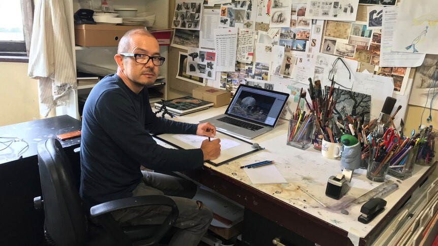 A man with glasses sits at a desk with tubs of pencils and brushes and lots of pictures stuck on the wall. 