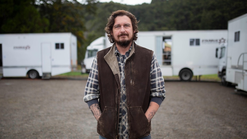 A man standing in front of film trailers looks seriously at the camera.