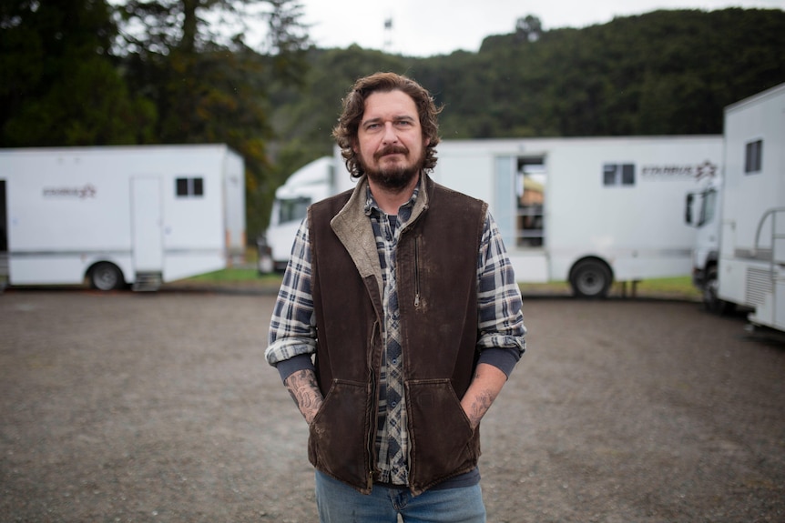 A man standing in front of film trailers looks seriously at the camera.