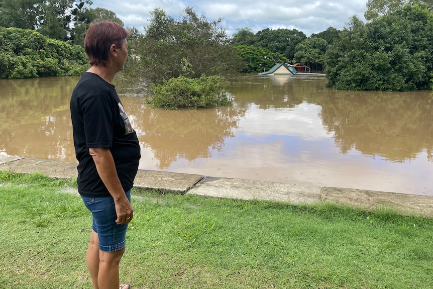 A woman in shorts and a T-shirt looks across flood waters to her partially submerged house.