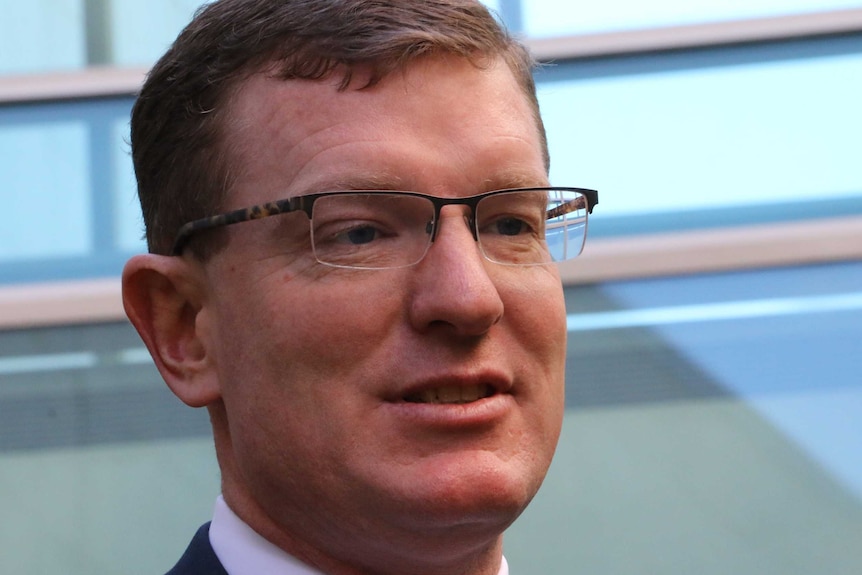 A bespectacled man with short, dark hair, wearing a dark suit, light shirt, and tie.