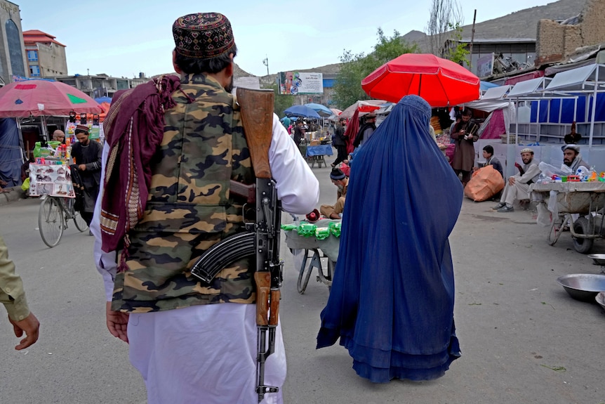 From behind, an Afghan woman wearing a burka is followed by a Taliban patrol guard with a gun.