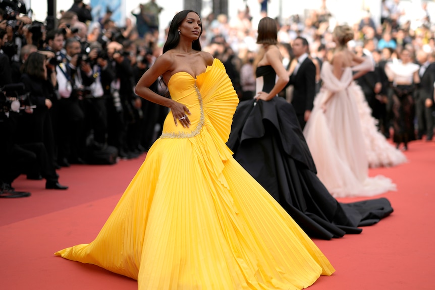 Jasmine Tookes wearing a strapless yellow gown with a full pleated skirt and on pleated fan detail on the side of the bodice. 