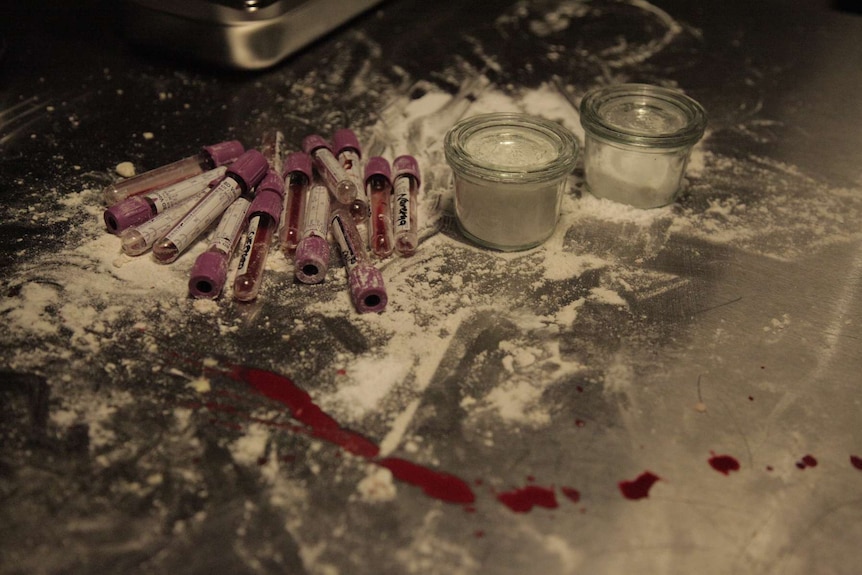 Stainless steel kitchen surface with two small cannisters of flower and sugar, and scattered medical vials with blood.