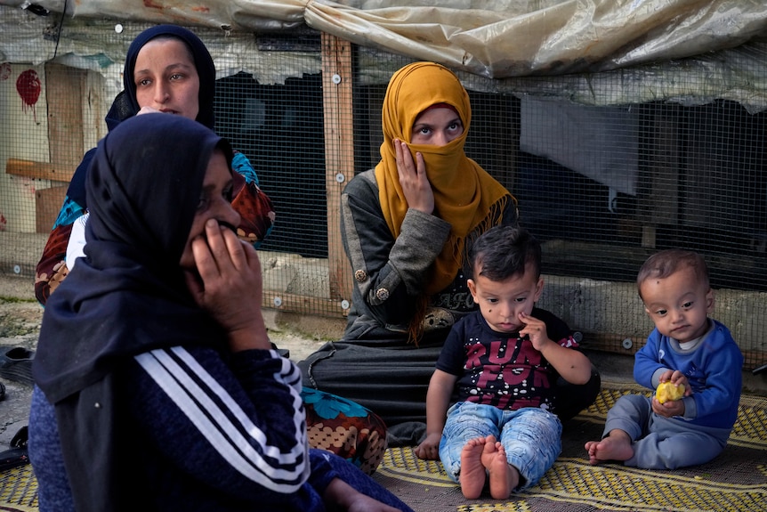 Three women and headscarves sit with two infant children. 