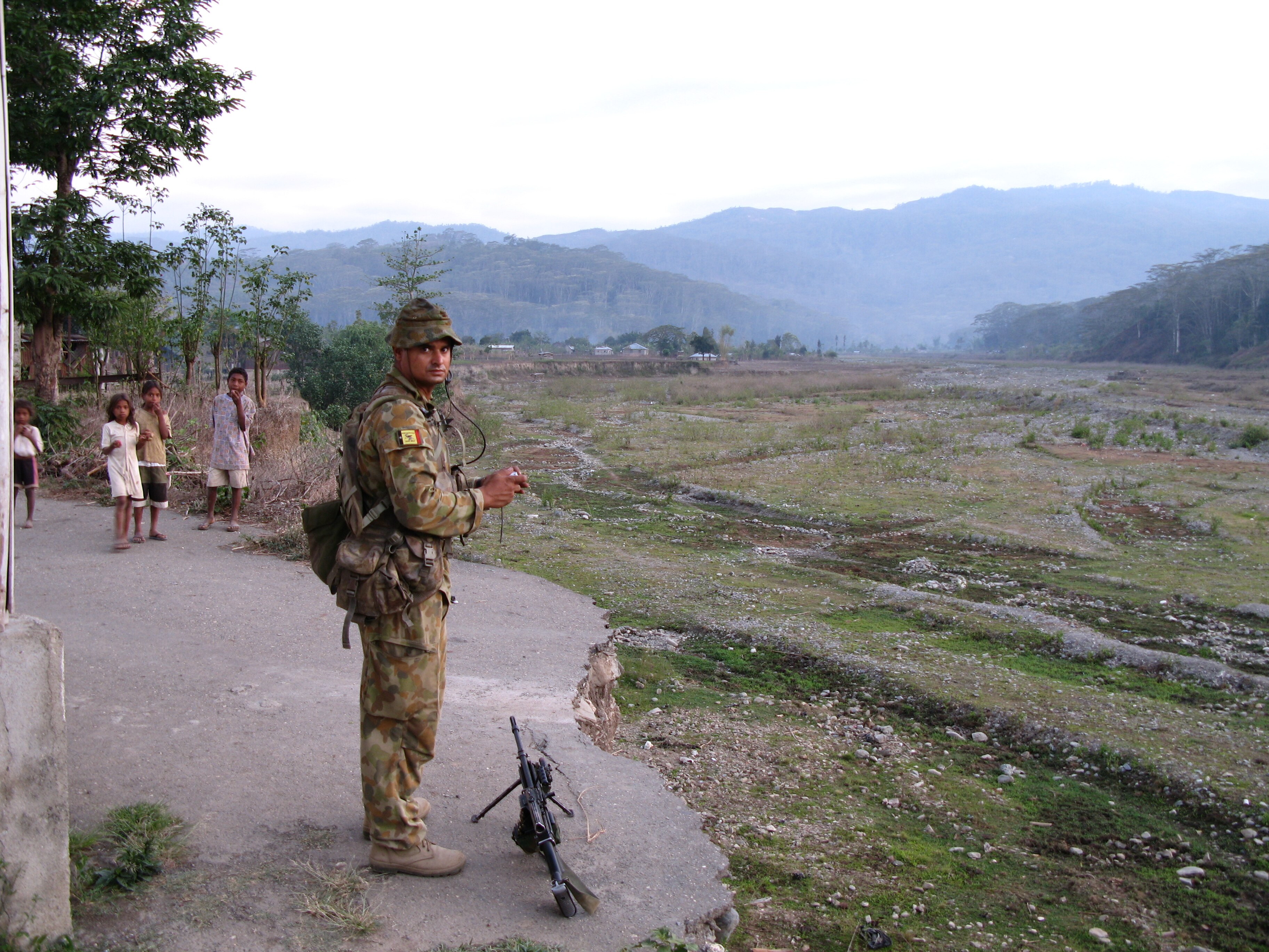 Michael Jefferies standing outside wearing a military uniform stands with his gun at his feet with some children behind him.  
