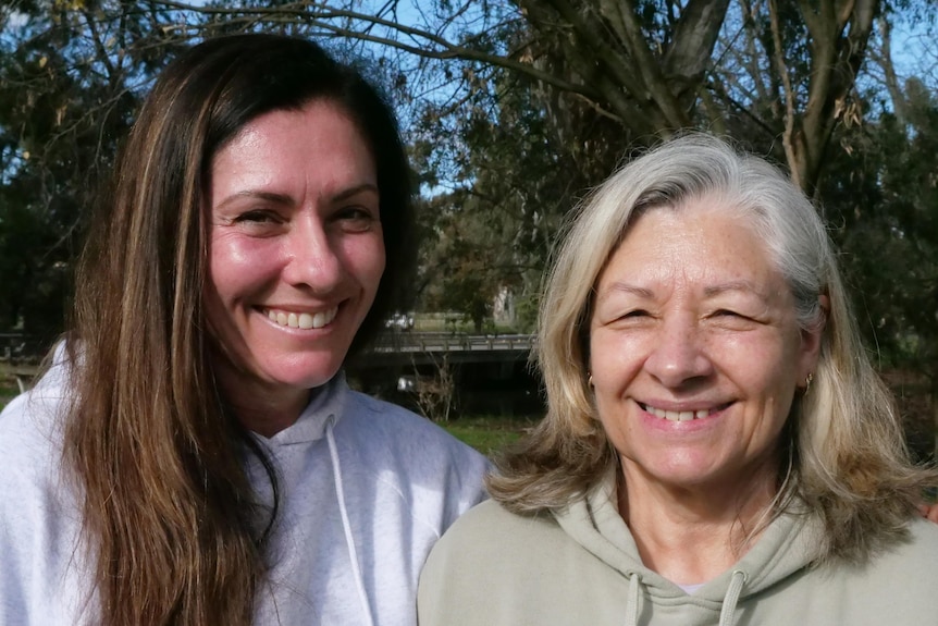 Two woman, one with white hair and one with long brown hair stand in front of a bridge and some trees.