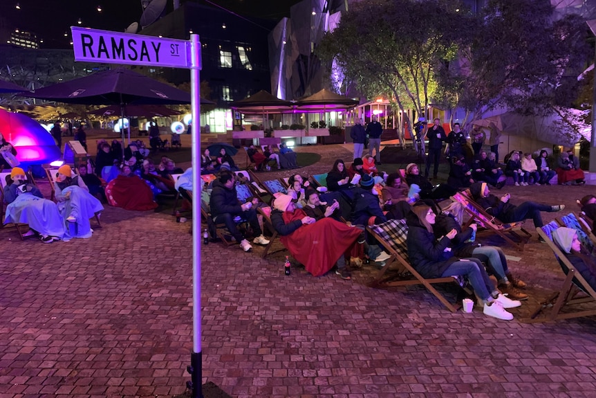 a group of people sitting on chairs watching a screen above them.