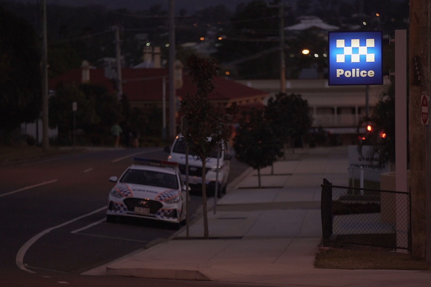 a blue sign with white writing that says police illuminates on an evening outside a police station. Two police vehicles parked