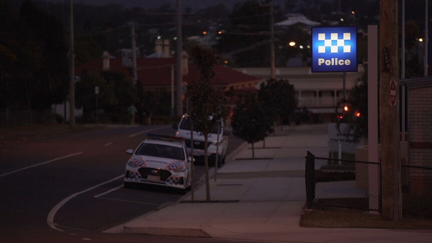 a blue sign with white writing that says police illuminates on an evening outside a police station. Two police vehicles parked