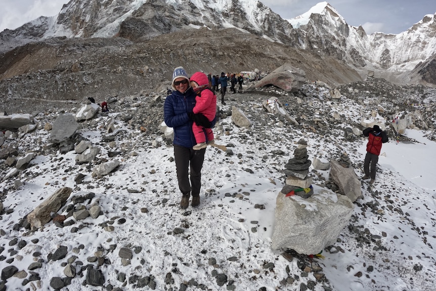 A  smiling woman holds her little daughter in a snowsuit as they stand among snowy mountains.