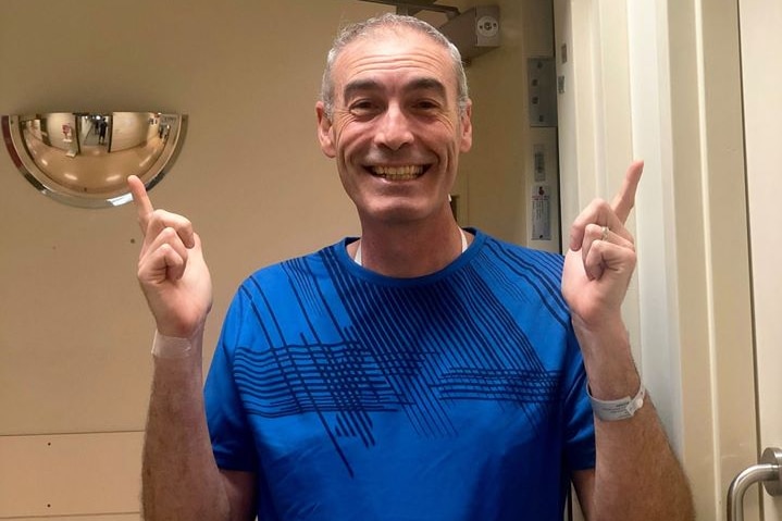 Greg Page points his fingers upwards towards an exit sign as he wears a blue shirt with a hospital wristband on. He is smiling.