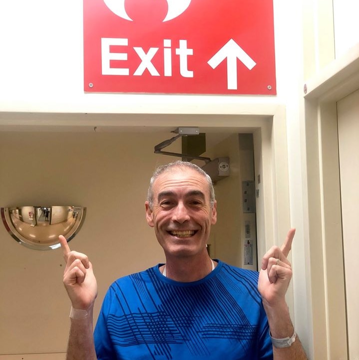 Greg Page points his fingers upwards towards an exit sign as he wears a blue shirt with a hospital wristband on. He is smiling.