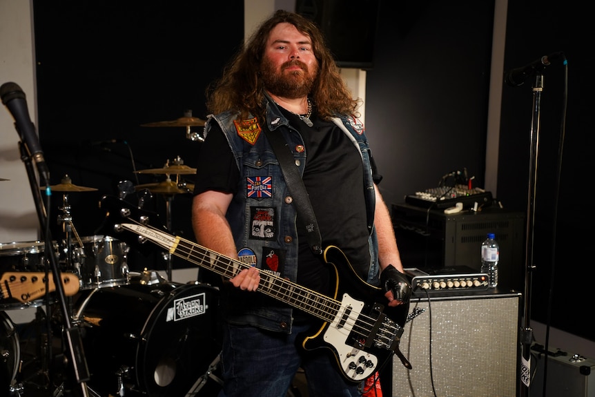 A rock guitarist with long hair and a beard stands posing for a photo holding his guitar in front of band equipment.
