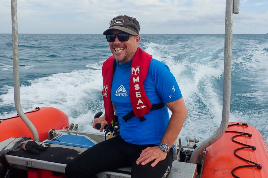 A man in a blue rash shirt, visor, black sunglasses sit on the back of an orange boat tender in the middle of the ocean.