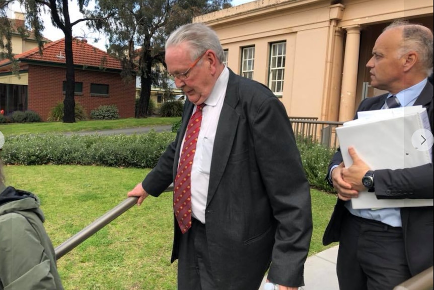 An older man dressed in a dark suit walks away from a courthouse.