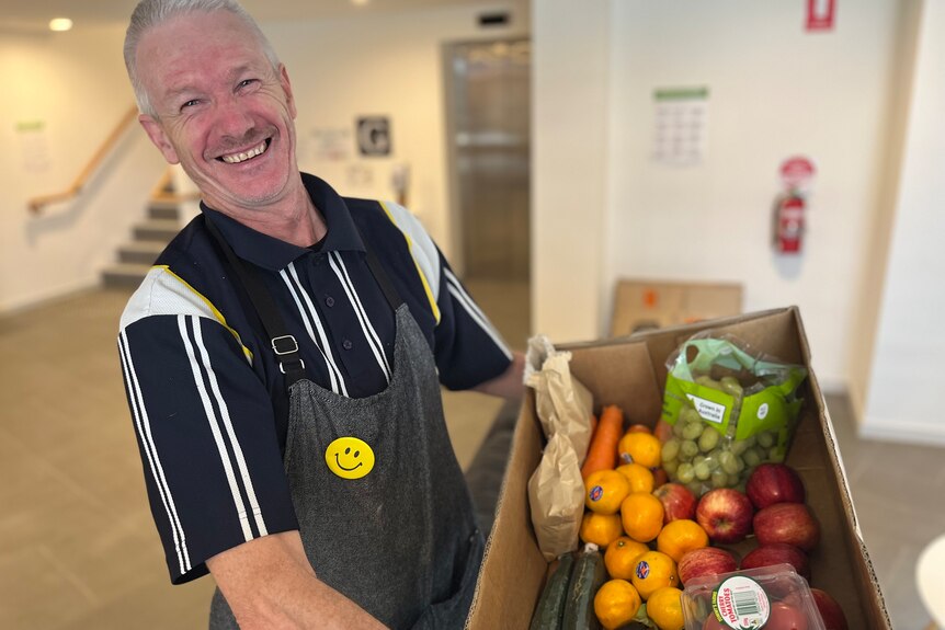 A smiling man in an apron holds a cardboard box full of fruit and vegetables
