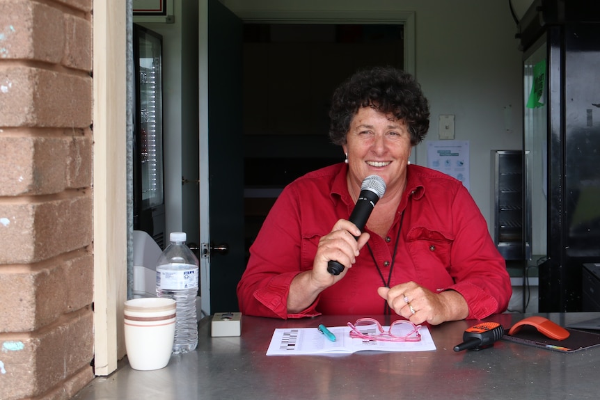 Smiling woman in red shirt with curly hair holding a microphone from a country football club canteen window