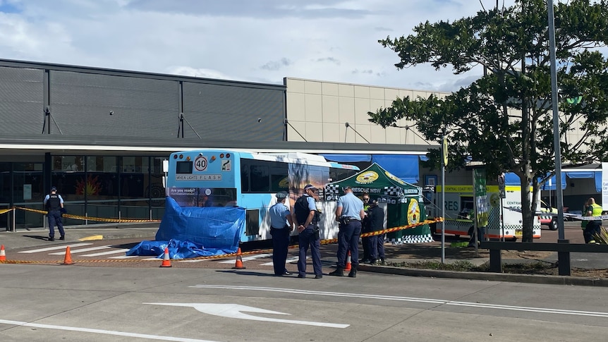 wide photo of a blue bus sitting idle in front of a shopping centre. Surrounded by blue tarp and blue and white police tape