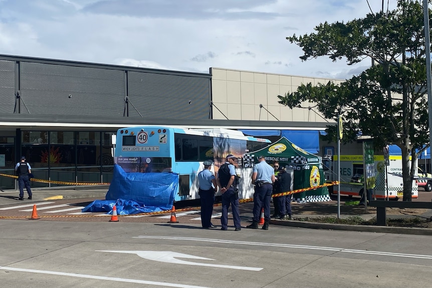 Police officers stand near a bus with a tarpaulin hanging off it in a shopping centre car park.