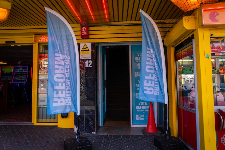 Two flags promoting Reform UK fly in front of an open door in the middle of an amusement park