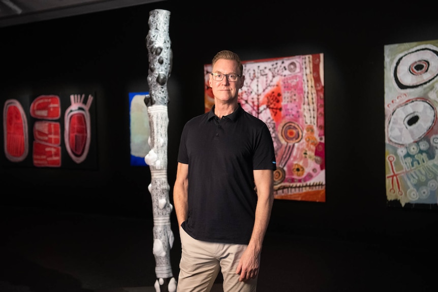 A man in a black t-shirt stands in front of several works of Indigenous art, bright and bold in colour. The background is black.
