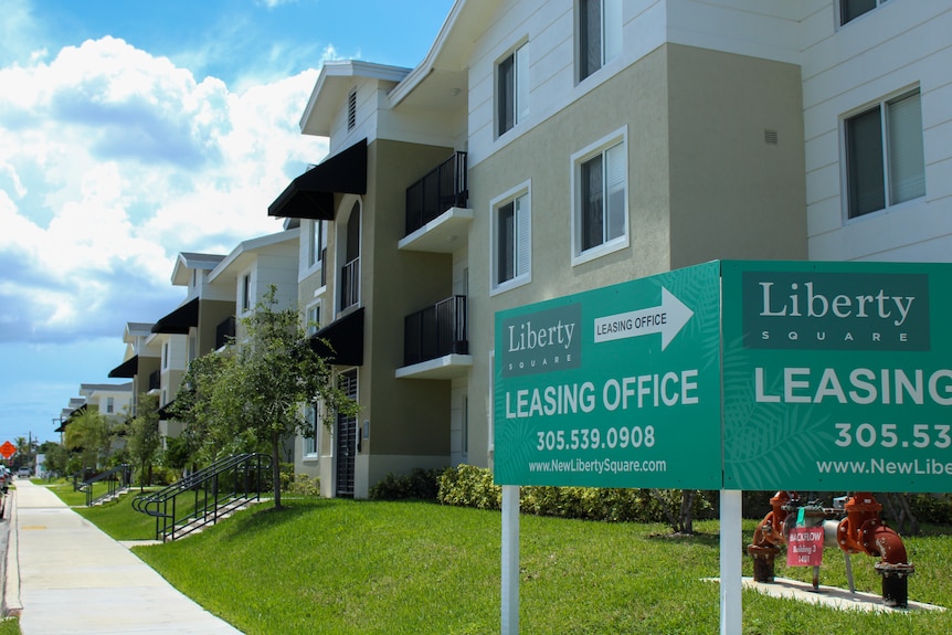 A three-story apartment building with black awnings bears a "now leasing" sign in front.