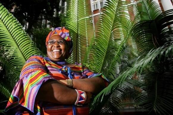 A middle-aged African woman in bright clothing stands with her arms crossed in front of a fern.