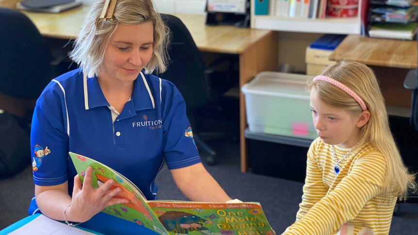 A woman in a blue shirt with an open book, reading to a blonde haired young girl with a yellow dress