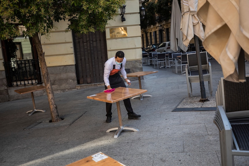 A waiter wearing a face mask wipes down an empty table as he disinfects an alfresco area.