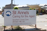 A sign that reads "St Anne's Caring for the Aged" outside a large building beneath a sunny sky.