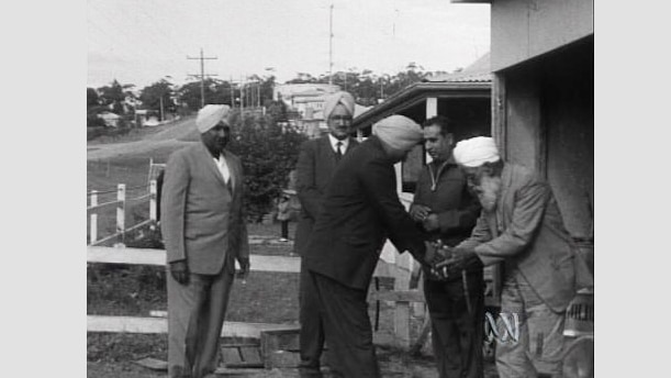 A group of Sikh men in suits and turbans stand in a house front yard, two men shake hands