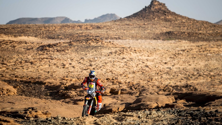 A motorcyclist rides up a rocky hill with the desert in the background in the Dakar Rally.