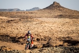 A motorcyclist rides up a rocky hill with the desert in the background in the Dakar Rally.