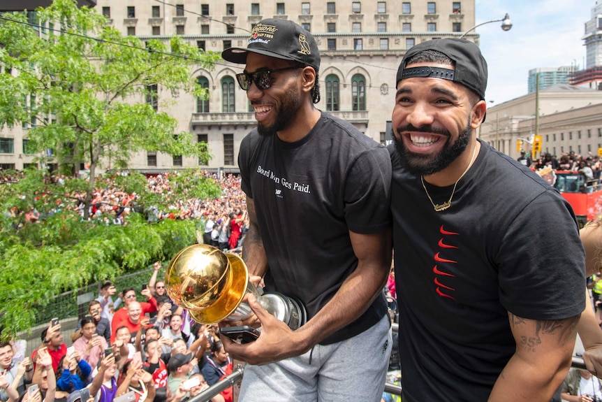 Kawhi Leonard and Drake stand on a platform above a crowd, Leonard holding a golden trophy.
