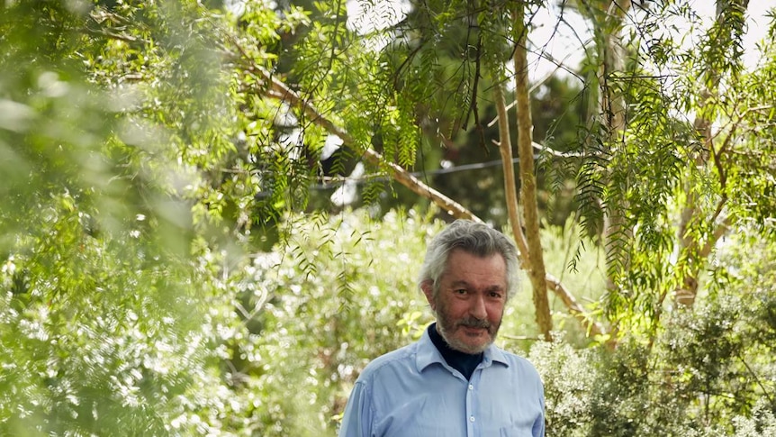 Robert Manne in blue tshirt and jeans standing in a green, leafy garden, next to a dog.